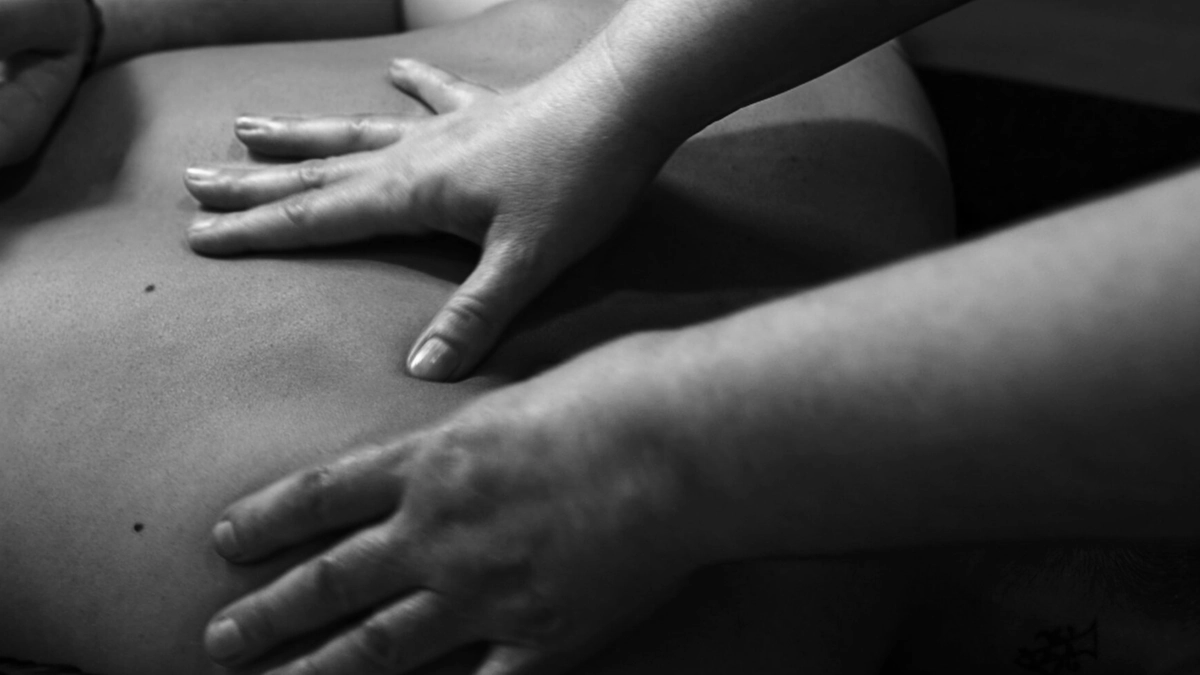 Two hands pressing along a client's back in near-darkness, photographed in black and white