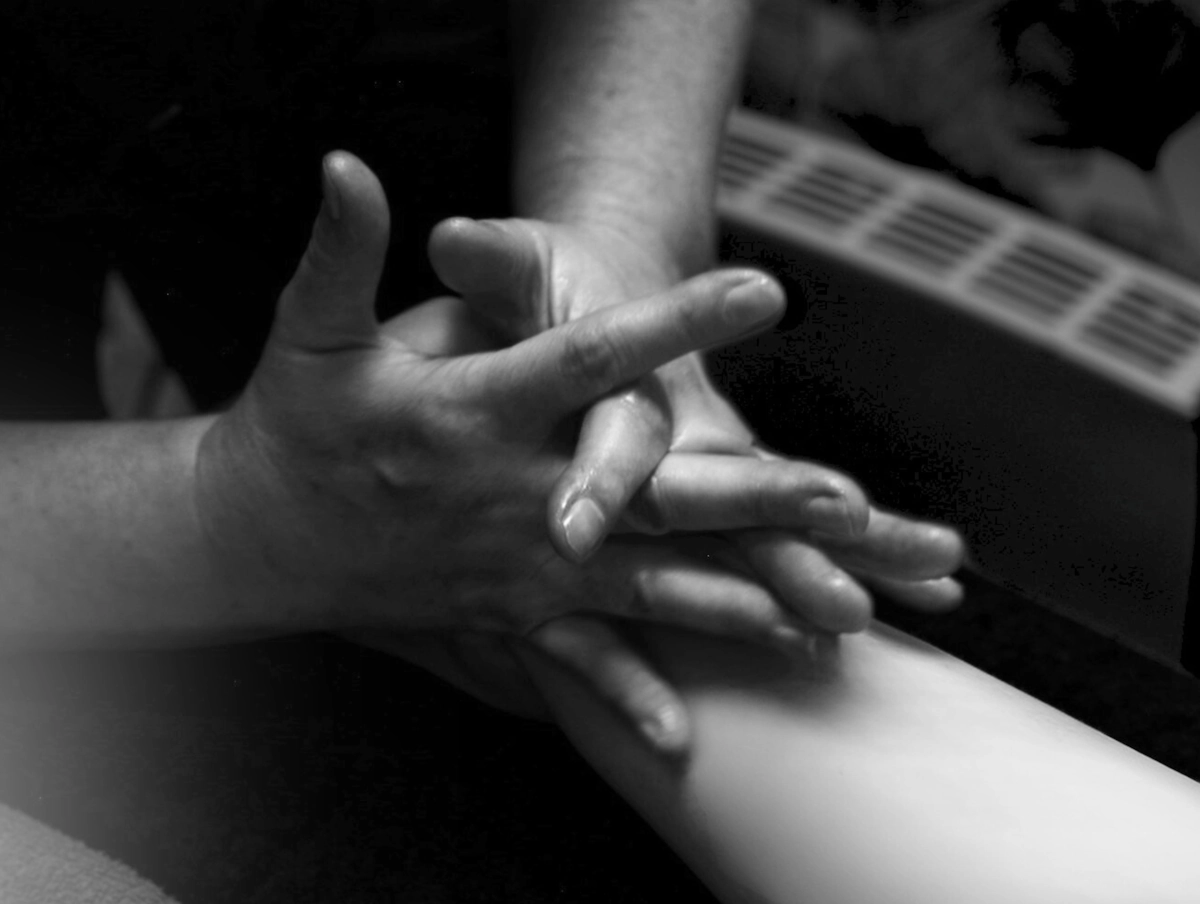 Close black-and-white photograph of Sylvie's hands interlaced with a client's forearm during a massage