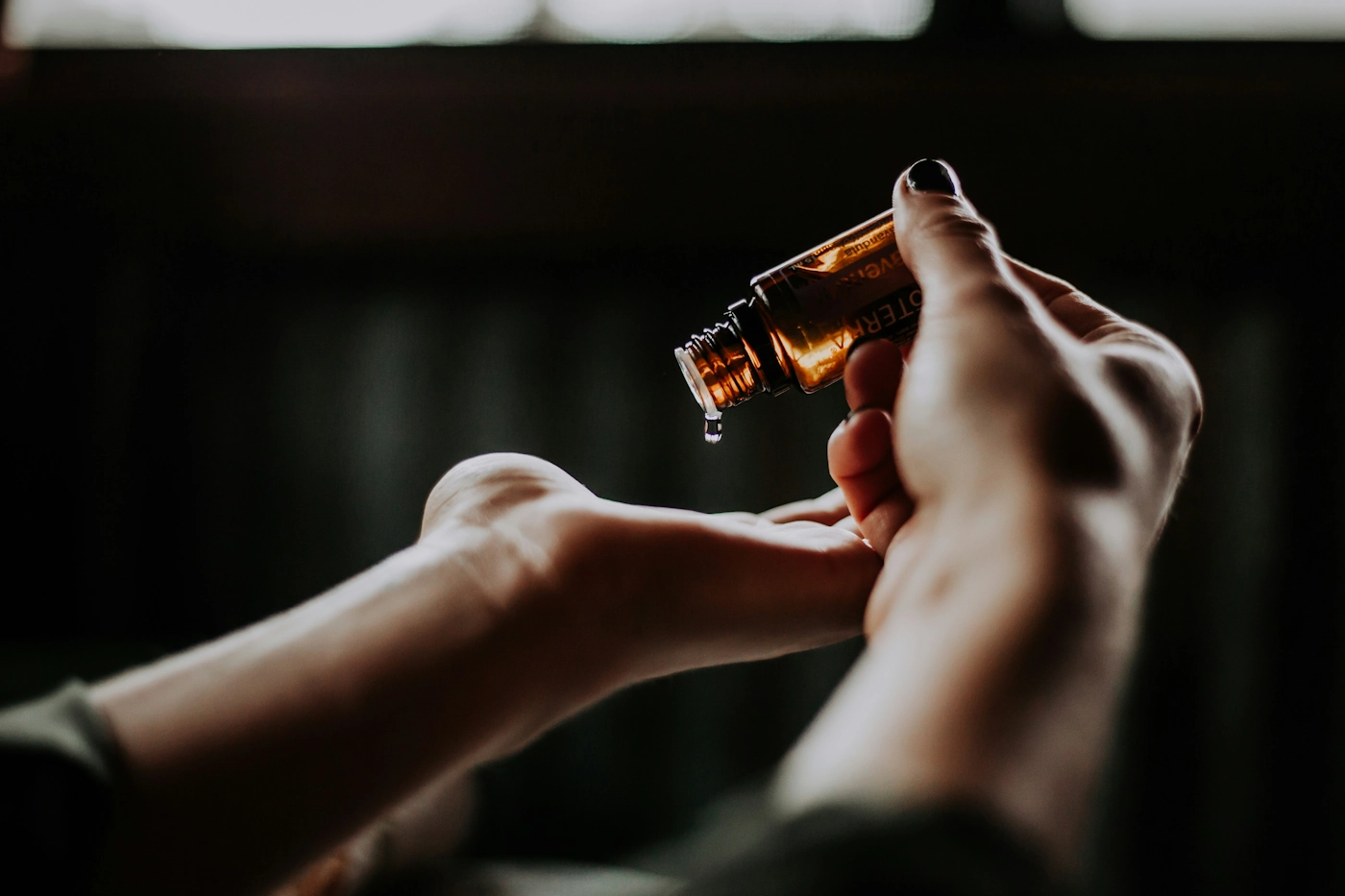 An amber bottle of massage oil tipped over Sylvie's palm, a single drop falling, lit warm against a dark room